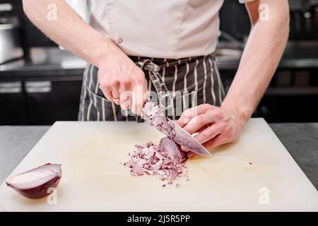 Close-up of cook in apron using professional knife and cutting onion on cutting board at table Stock Photo