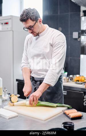 Young chef in apron cutting celery on cutting board to prepare dish with fish at table in kitchen Stock Photo