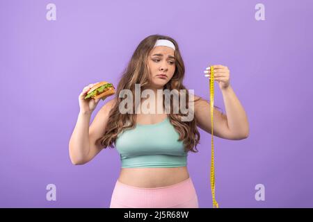 Sad unhappy hungry young european plus size lady in sportswear with burger in hand looks at measuring tape Stock Photo