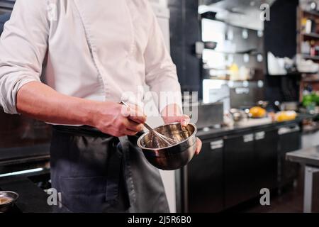 Close-up of chef in apron whipping eggs in bowl with a whisk while cooking in kitchen Stock Photo
