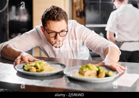 Serious chef in eyeglasses looking at plates with main course on the counter and preparing them for delivery Stock Photo