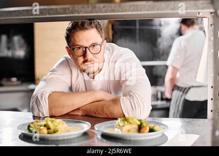Portrait of young professional cook in eyeglasses looking at camera behind the counter with ready dishes Stock Photo