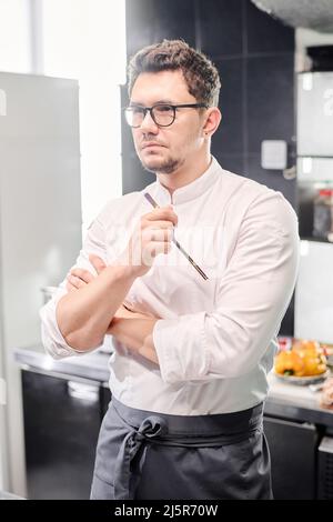 Young serious chef in eyeglasses standing in uniform in kitchen and thinking over new menu Stock Photo
