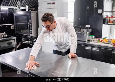 Young professional cook in uniform wiping kitchen counter in kitchen after cooking Stock Photo