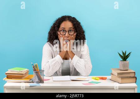 Shocked young black woman in eyeglasses sitting at table with study materials on blue studio background Stock Photo