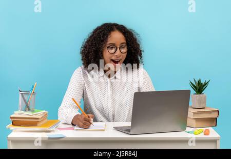 Online education concept. Excited young black female student sitting at table, using laptop, writing during web lesson Stock Photo