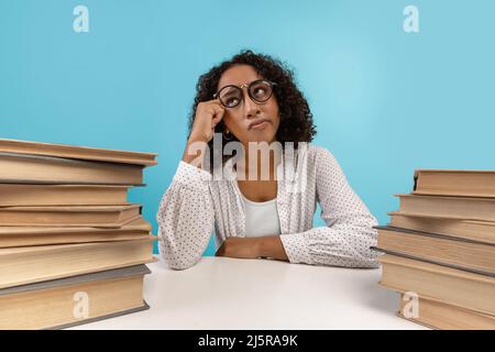 Young black female student in glasses sitting at desk with piles of books, daydreaming, feeling bored during lesson Stock Photo