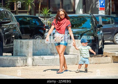 Mother and little son walk on street of Omis against parked on lot cars on sunny day. Woman and toddler boy enjoy summer vacation in Croatia Stock Photo