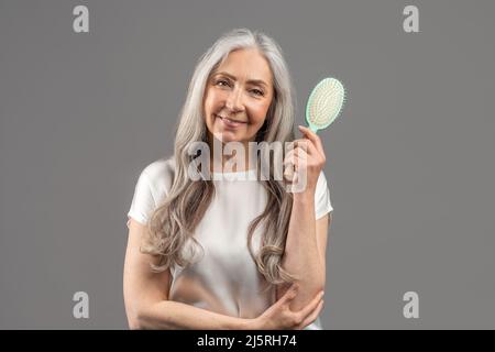 Portrait of lovely senior lady with beautiful long hair holding brush and smiling at camera on grey studio background Stock Photo