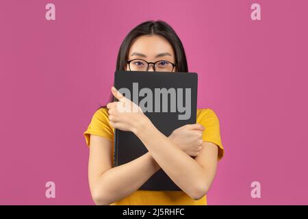 Asian female student in glasses hiding behind laptop, studying for college exams online, standing on violet background Stock Photo