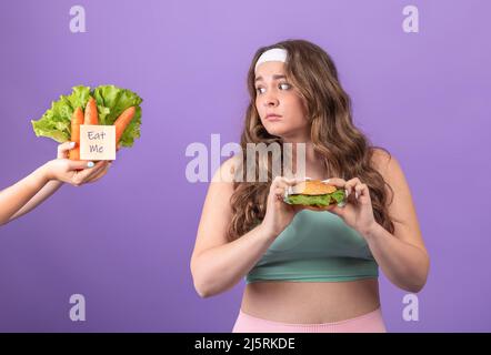 Sad hungry young european plus size woman in sportswear with burger in hands looks at carrot and salad Stock Photo