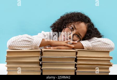 Sleepy young black female student leaning on big stacks of books, tired of getting ready for exam, feeling bored Stock Photo