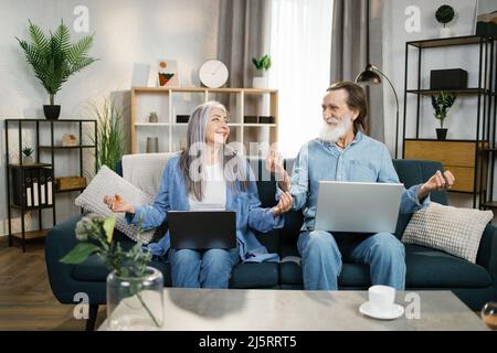 Smiling aged couple pensioners spouses spending time together, wife and husband sitting in lotus position in light comfortable living room. Old people care of physical and mental health concept Stock Photo