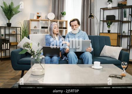 Mature man and woman using laptops, looking at screen, older spouses browsing apps, shopping or chatting online, making video call, using bank service, sitting on cozy sofa in living room Stock Photo