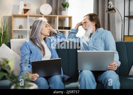 Positive attractive mature aged spouses spending weekend together sitting on comfortable couch in living room at modern cozy country home holding on lap computer watching online movie having good mood Stock Photo