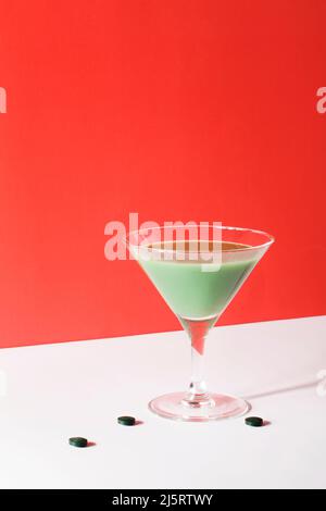 green smoothie in martini cocktail glass with spirulina algae pills on red background. self care and minimal healthy party life concept. copy space Stock Photo