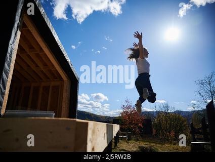 Happy woman on construction site jumping from terrace at unfinished wooden frame house on sunny day. Stock Photo