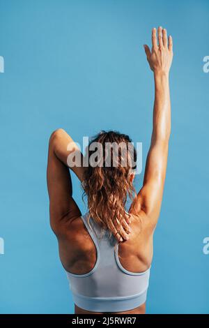 Rear view of young muscular woman stretching her arm on blue background Stock Photo