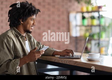 Happy young african american curly man with smartphone typing on laptop, sits at table with cup Stock Photo