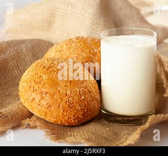 Still life with a glass of milk and two buns. A glass of homemade milk and two appetizing freshly baked sesame buns are placed on a coarse fabric. Stock Photo