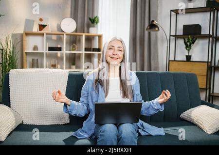 Relaxed pretty grey-haired lady sitting on couch with laptop on knees and meditating with closed eyes. Tired cheerful mature woman freelancer taking break during working process at home. Stock Photo