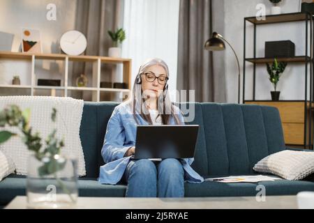 Beautiful senior woman using headset and laptop for video conversation at living room at home. Pleasant mature lady female freelancer sitting at table and taking notes during video chat. Stock Photo