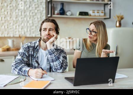 Pensive sad young european husband with stubble and wife in glasses pay bills and taxes at table with laptop Stock Photo
