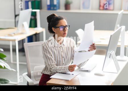 Concentrated latin businesswoman working with documents and reports, sitting at workplace in office interior Stock Photo