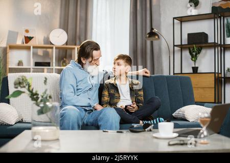 Happy grandfather with grey beard cuddling smiling small child boy playing online mobile game together at home. Joyful different generations family using funny smartphone applications, resting on sofa Stock Photo