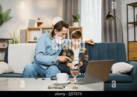 Happy senior grandfather and cute grandson using smart phone apps at home, old grandpa embracing grandchild holding looking at cellphone screen playing mobile game relaxing sit on sofa Stock Photo