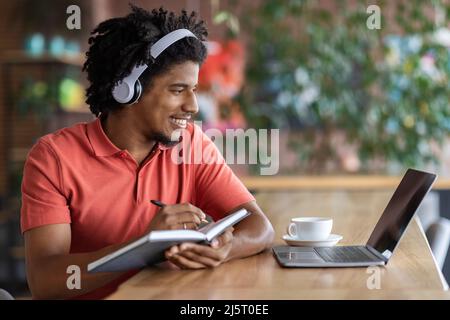 Smiling young black curly man in wireless headphones makes notes in notebook and looks at laptop in cafe Stock Photo