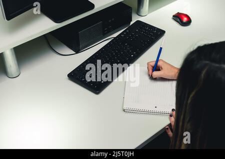 unrecognizable young woman sitting in her office writing in pad, with keyboard and computer cassette on desk, copy space Stock Photo
