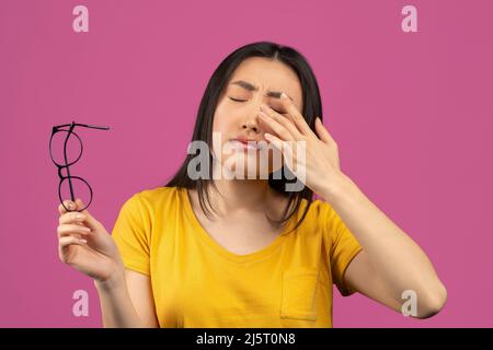 Young asian lady suffering from eye pain, taking off glasses and touching her eye, standing over violet background Stock Photo