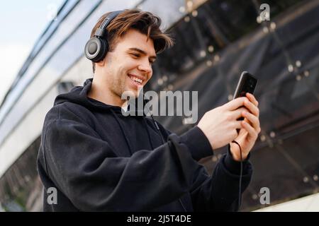 Young Caucasian Man In Headphones Using Smartphone Texting And Listening To Music. Stock Photo