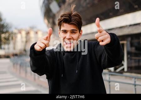Portrait of a cheerful young man in black hoodie pointing his finger at you on a urban background. Stock Photo