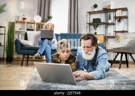 Happy different ages family leisure at home concept, mature grandmother relaxing using laptop on sofa in comfort living room while her grandchild playing on laptop with grandpa on warm floor. Stock Photo