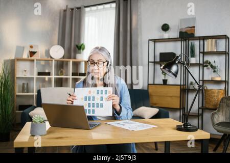 Cheerful mature woman presenting financial report to colleagues through video call on laptop. Positive older woman freelancer sitting at workplace and talking in headset. Stock Photo