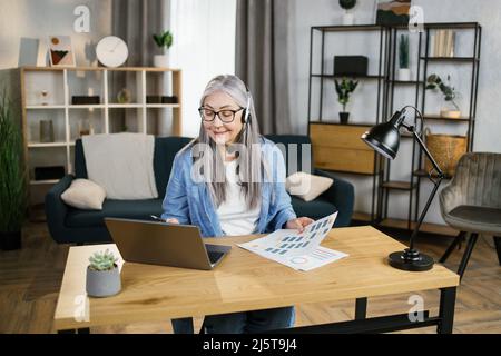Cheerful mature woman presenting financial report to colleagues through video call on laptop. Positive older woman freelancer sitting at workplace and talking in headset. Stock Photo