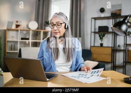 Cheerful mature woman presenting financial report to colleagues through video call on laptop. Positive older woman freelancer sitting at workplace and talking in headset. Stock Photo