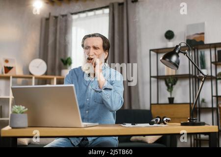 Elegantly looking aged grey-beard mature man chief of big company relaxing at office smoking cigar while gazing at the screen of laptop while sitting at the tabe. Stock Photo