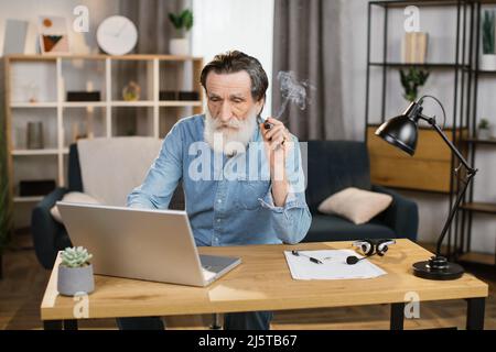 Elegantly looking aged grey-beard mature man chief of big company relaxing at office smoking cigar while gazing at the screen of laptop while sitting at the tabe. Stock Photo