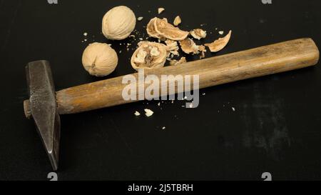 Three walnuts (one of them cracked) and a hammer on a dark scratched background Stock Photo