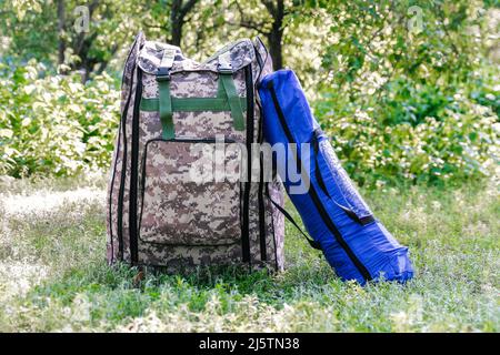 Defocus military backpack and blue tent or sleeping bag. Army bag on green grass background near tree. Military camouflage army rucksack. Tourist summ Stock Photo