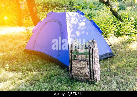 Defocus blue open tourist tent standing on green nature background. Army backpack. Tourism concept. Summer vacation in forest, camping. Lifestyle. Sun Stock Photo