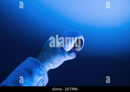 Blue color cast over male hand holding modern tiny 8k UHD surveillance camera with the cyclops like lens Stock Photo