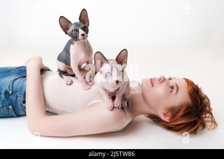 Two Canadian Sphynx Cat, sitting on young woman lying down on back on white background. Cute redhead woman with short hair wearing T-shirt and jeans Stock Photo