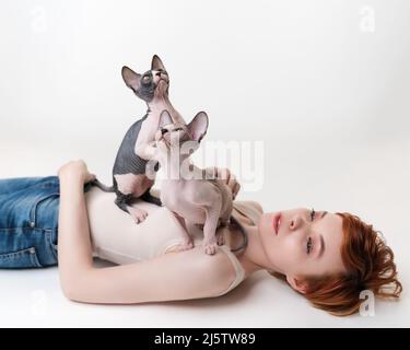 Two thoroughbred kittens looking up, sitting on redhead young woman lying on her back on white background. Pretty woman with short hair wearing T-shir Stock Photo