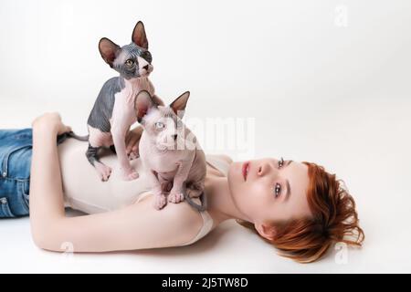 Two friendly Sphynx Hairless Cat, sitting on redhead young woman lying on back on white background. Beautiful woman with short hair Stock Photo