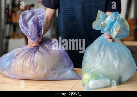 Two large cellophane sacks held by teenage guy standing by table in garage after sorting variety of waste into different bags Stock Photo