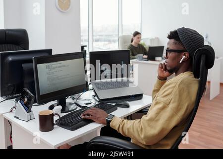 Side view of young serious lack man concentrating on decoding data while sitting in front of computer monitor by workplace in office Stock Photo